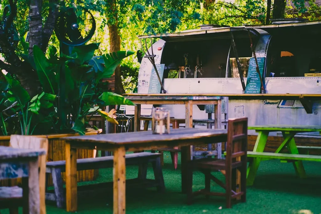 Outdoor food trailer dining area with wooden tables, turf, and foliage.