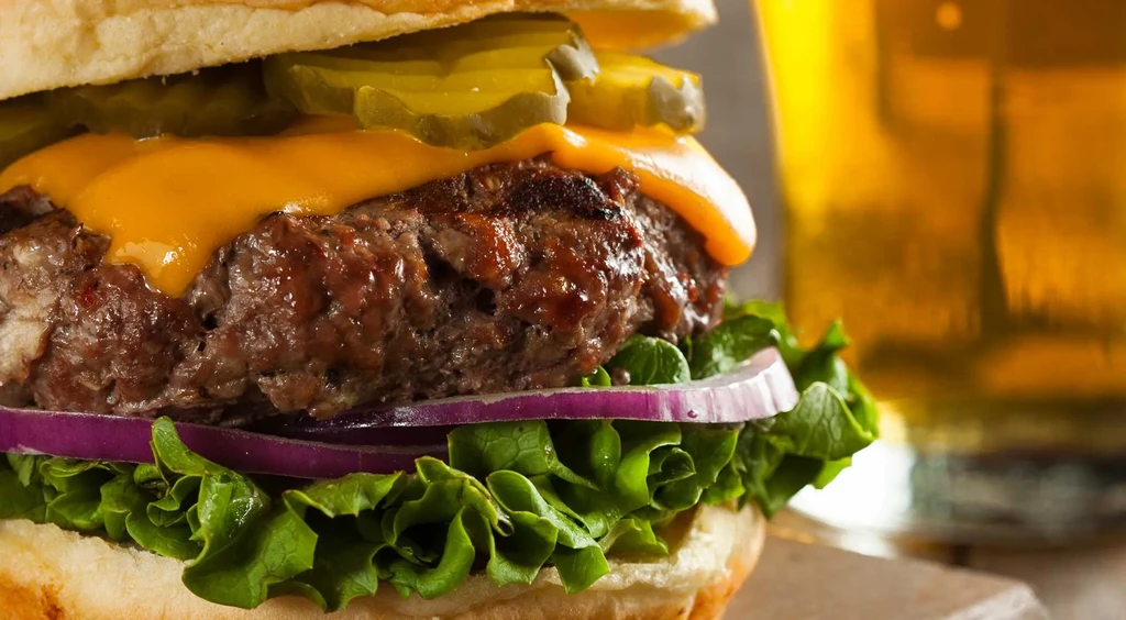Close-up cheeseburger with melted cheese, lettuce, onion, pickles, and beer.