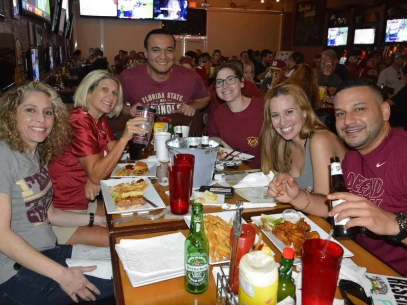 Six adults smiling, eating burgers and wings at a sports bar.