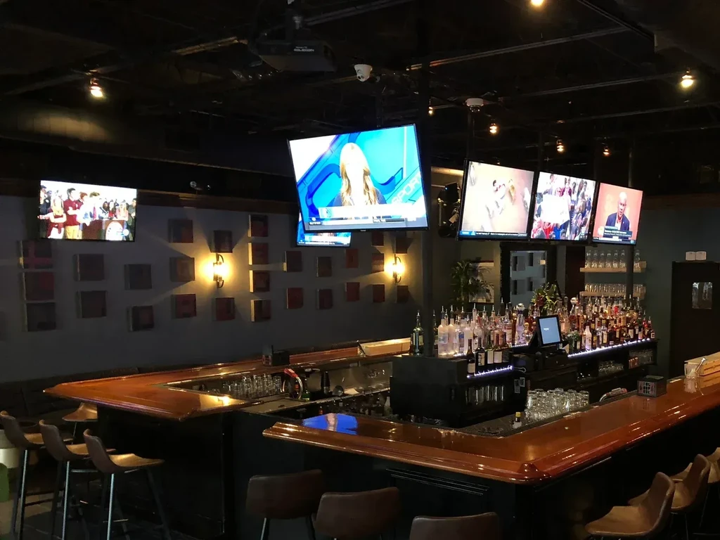 Dimly lit modern bar interior with counter, stools, and multiple TVs.