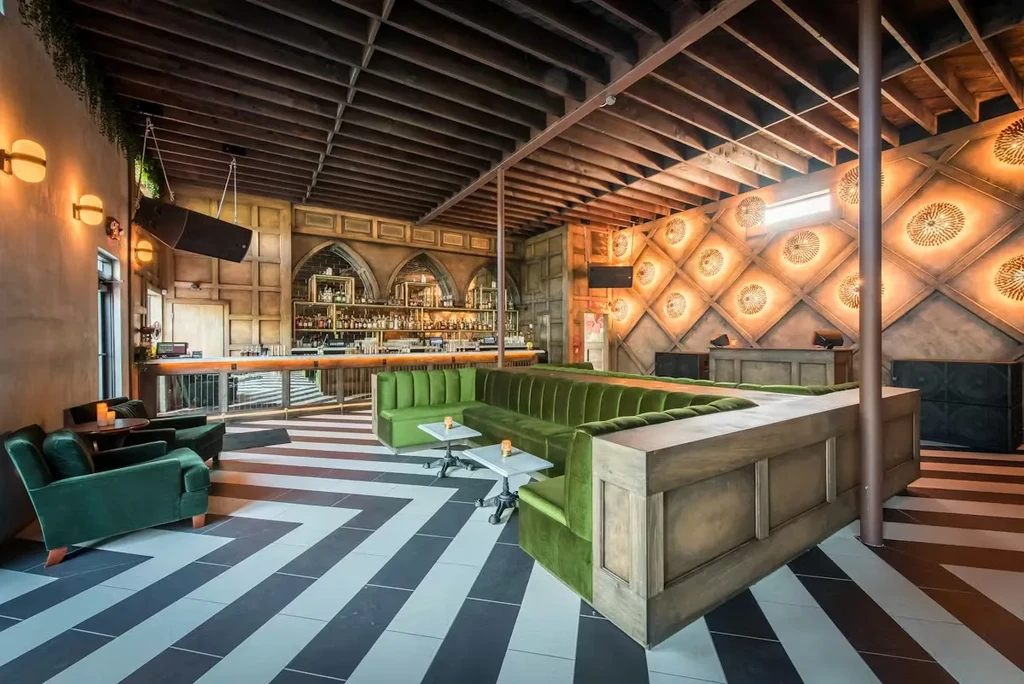 Stylish bar interior with wooden counter, chevron floor, and green velvet seating.