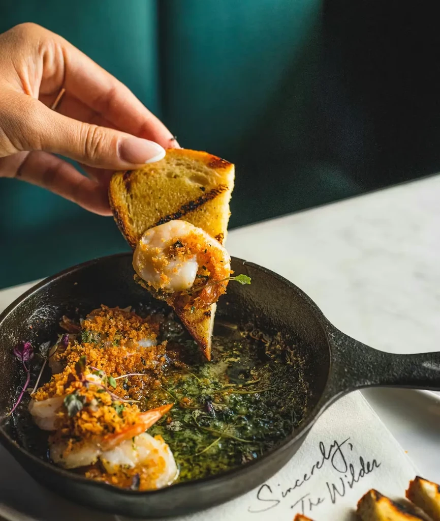 Hand scooping glistening garlic shrimp from a pan with toasted bread.