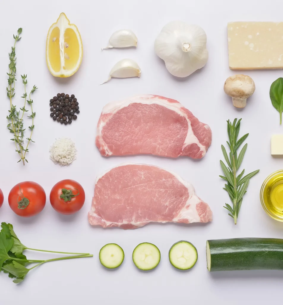 Flat lay of raw pork chops, vegetables, herbs, and seasoning ingredients.