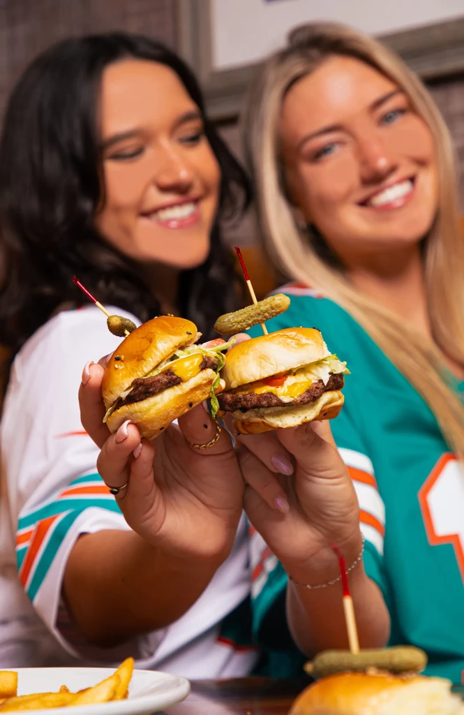 Two hands hold cheeseburger sliders with pickles; two smiling women blurred.