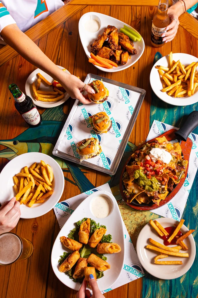 Hands sharing wings, nachos, sliders, fries, and beer at Quarterdeck.