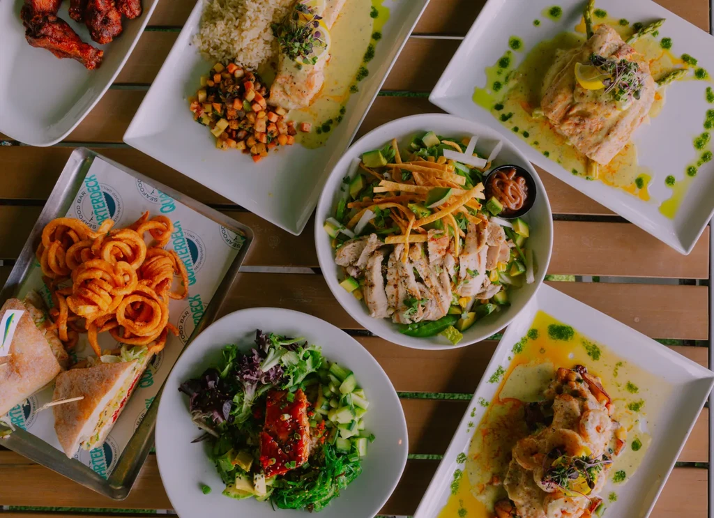 Overhead view: various grilled dishes, salads, sandwich, fries on table.