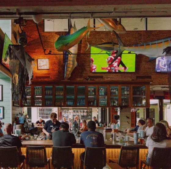 Bustling sports bar interior with patrons, bartender, fish, and TVs.