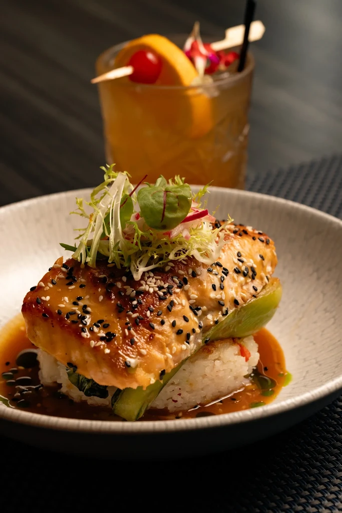 Close-up of sesame-crusted salmon with greens, rice, sauce, and a cocktail.