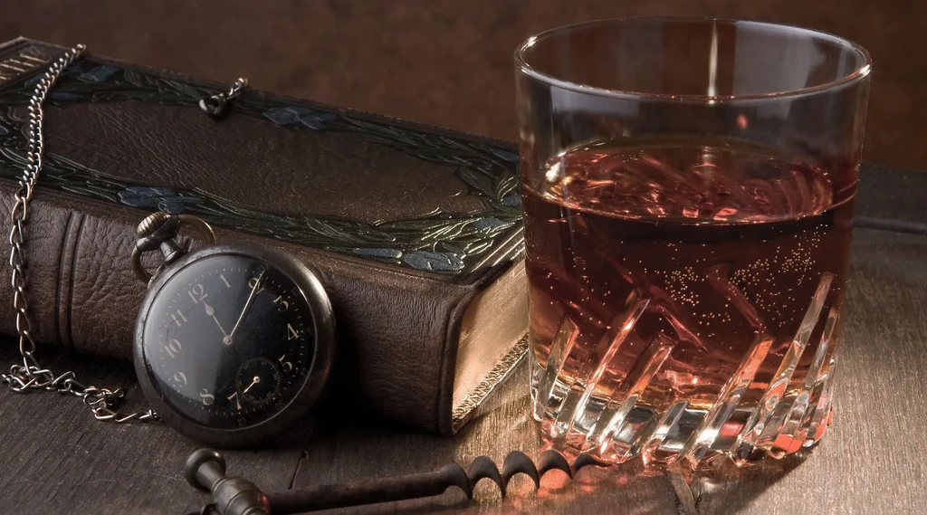 Whiskey glass, antique watch, and leather book on a dark surface.
