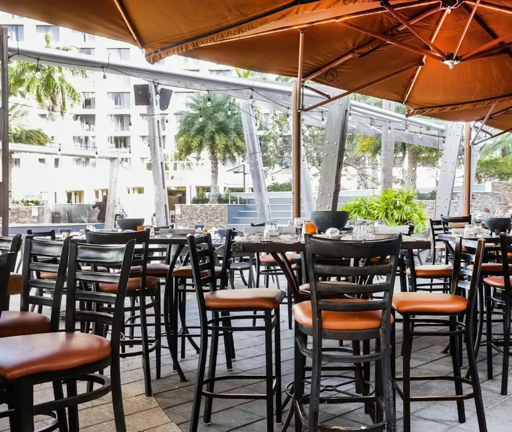 Modern outdoor patio with tall tables, bar stools, lit umbrellas, and palms.