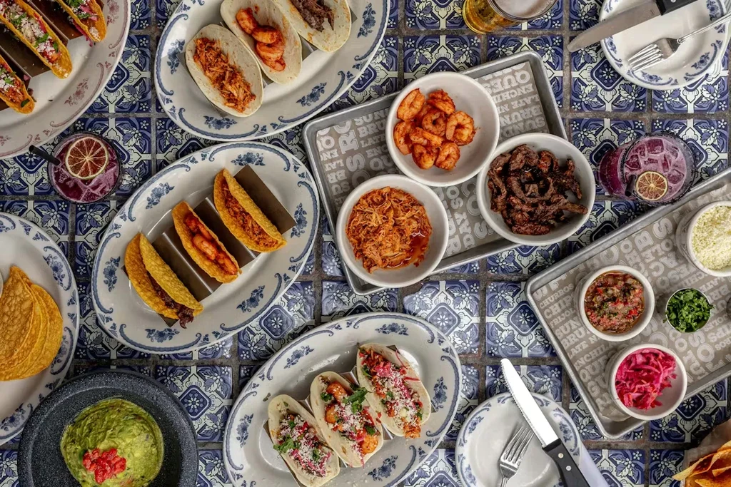 Overhead photo of Mexican tacos, toppings, and drinks on a blue tiled table.