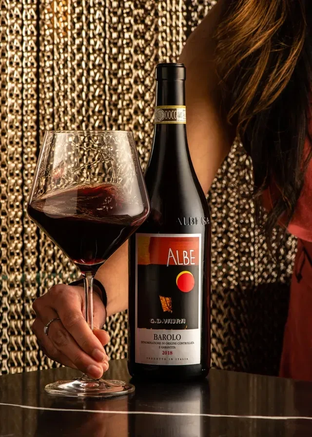 Albe Barolo wine bottle and glass, held by hand, golden background.