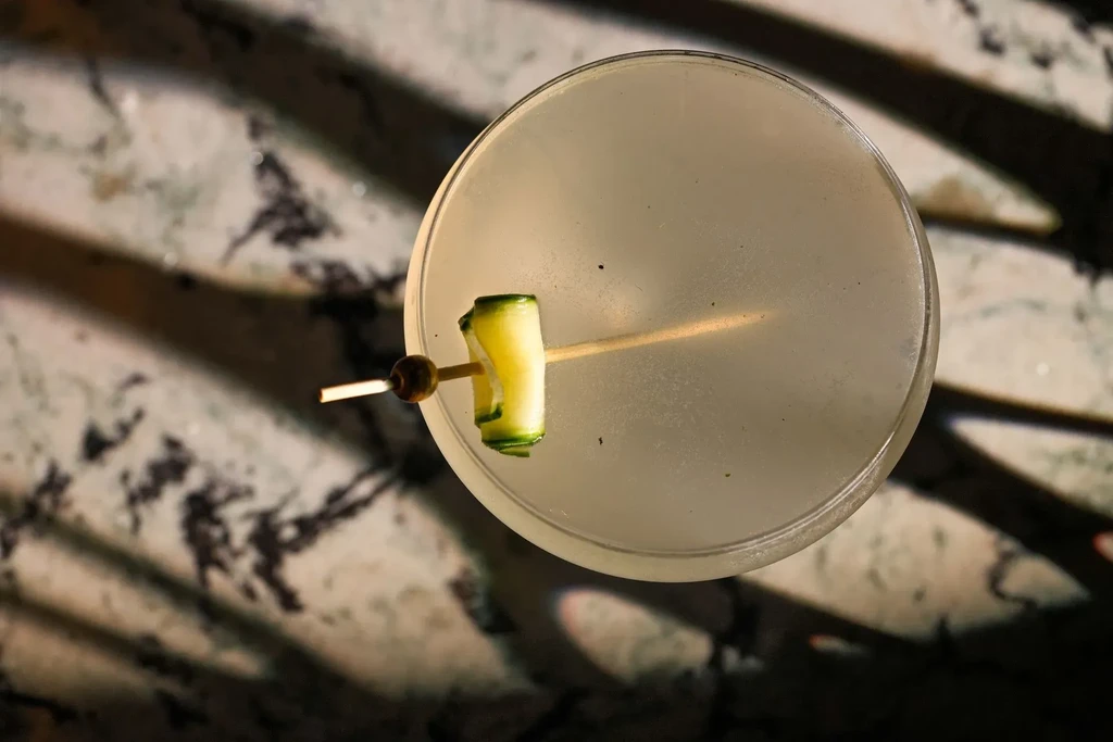 Overhead, pale cocktail in martini glass with rolled cucumber garnish.