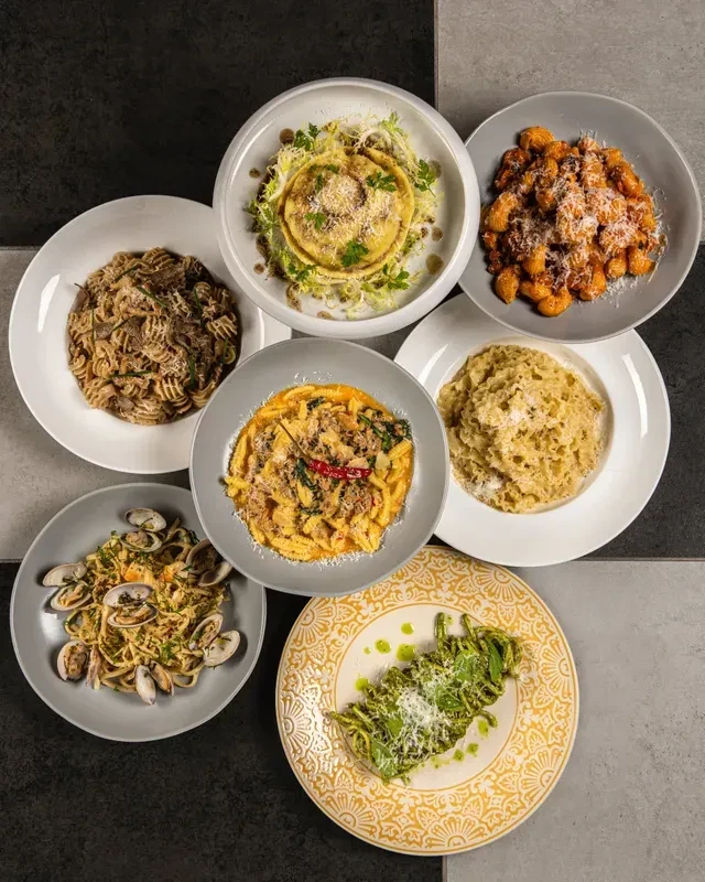 Overhead shot of seven distinct, colorful pasta dishes on a checkered surface.