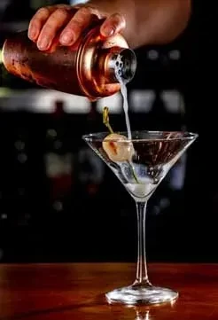 Bartender's hand pours beverage from copper shaker into garnished martini.