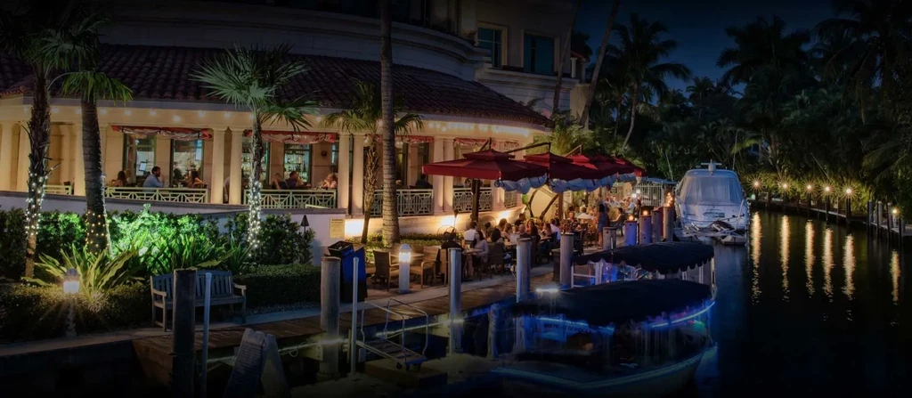 Lively waterfront restaurant with outdoor dining, red umbrellas, and boats at night.