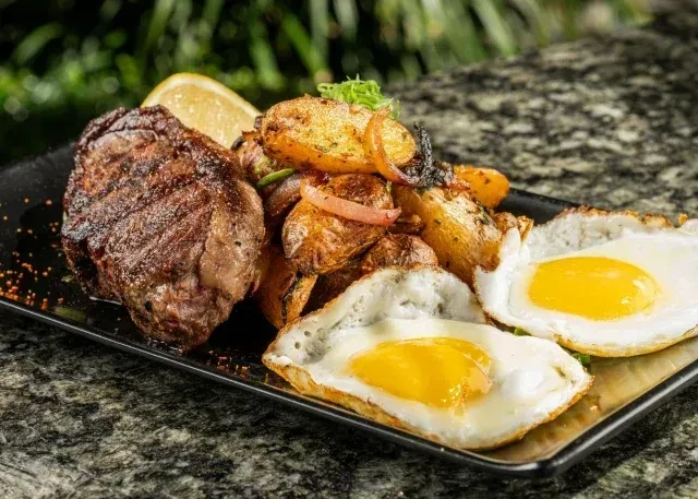 Grilled steak, roasted potatoes, two fried eggs, and lemon on dark plate.