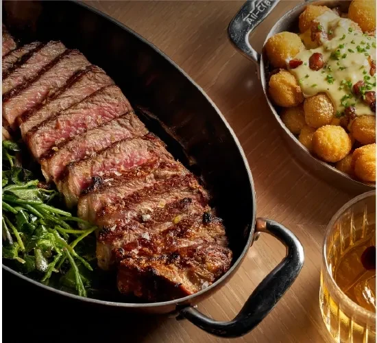 Sliced steak, potato croquettes with sauce, and a cocktail on a wooden table.