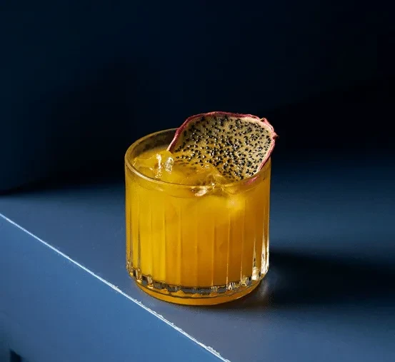 Yellow-orange cocktail with dragon fruit garnish in glass on dark blue.