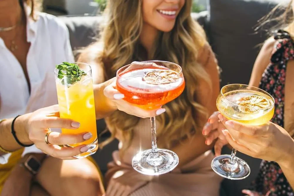 Three people's hands holding colorful, garnished cocktails; one woman smiles.