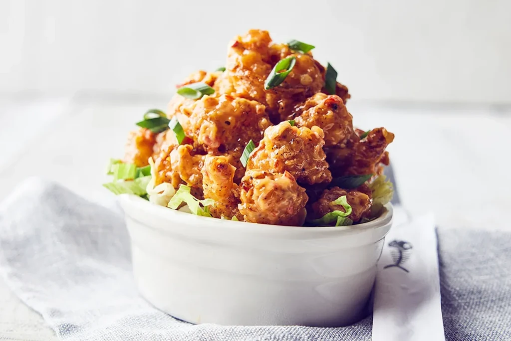 White bowl of crispy fried chicken/shrimp with orange sauce and green onions.
