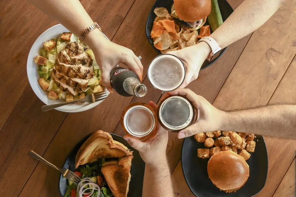 Overhead: four hands toast drinks over diverse plates of food.