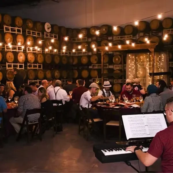 Rustic event space with barrels, dining guests, lights, and keyboard player.