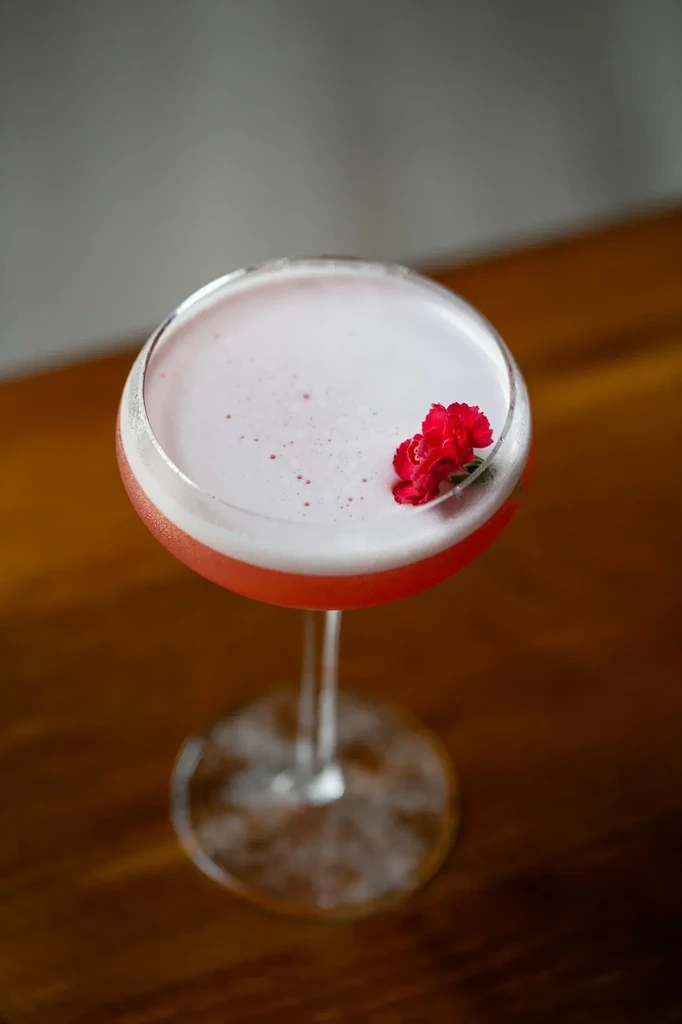 Red-pink cocktail in coupe glass with white foam and two red flowers.