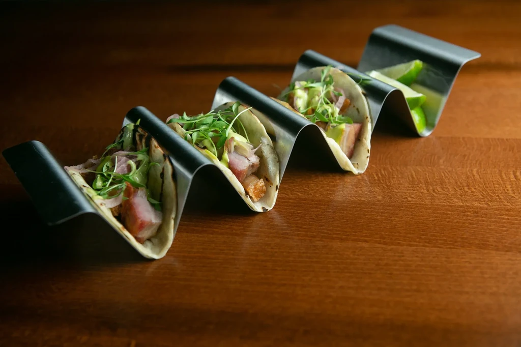 Four gourmet tacos with meat, red onion, green sauce, and lime wedges.