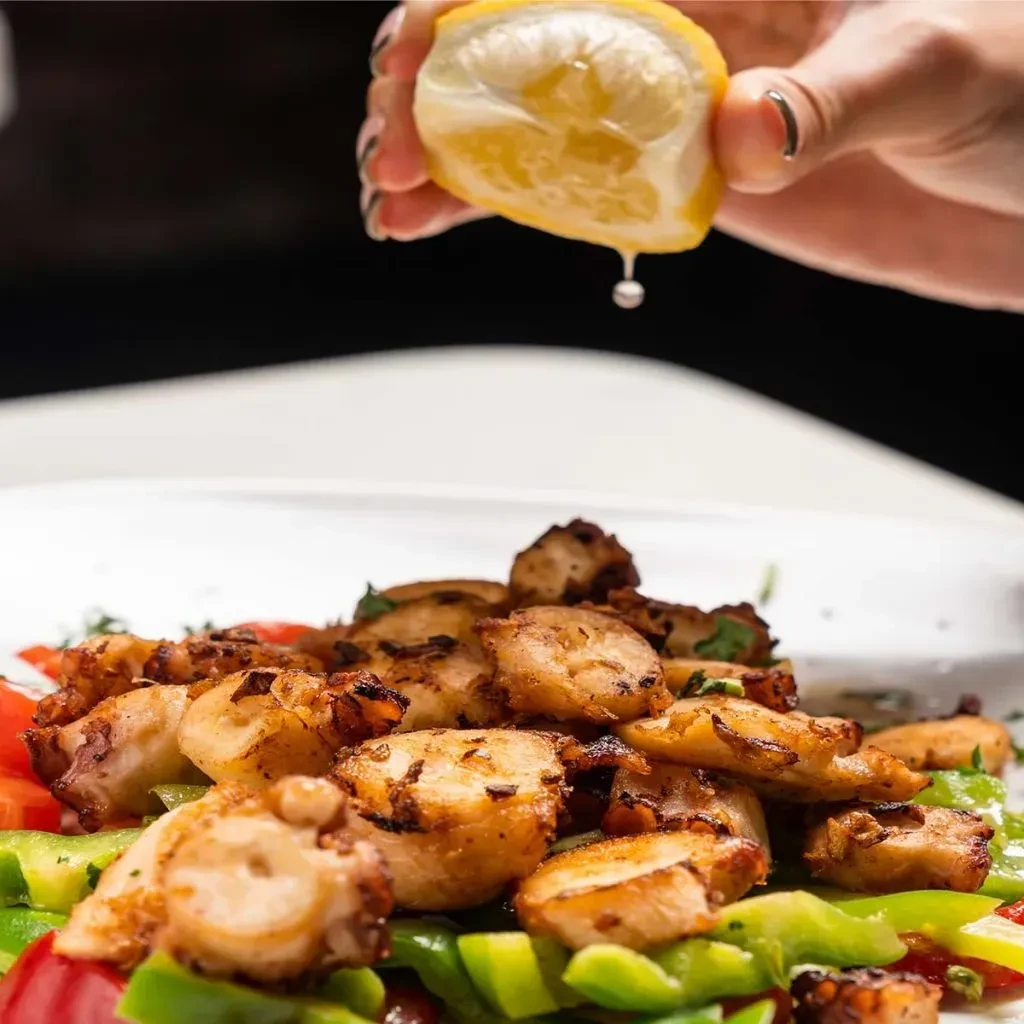 Grilled octopus with bell peppers and lemon being squeezed.