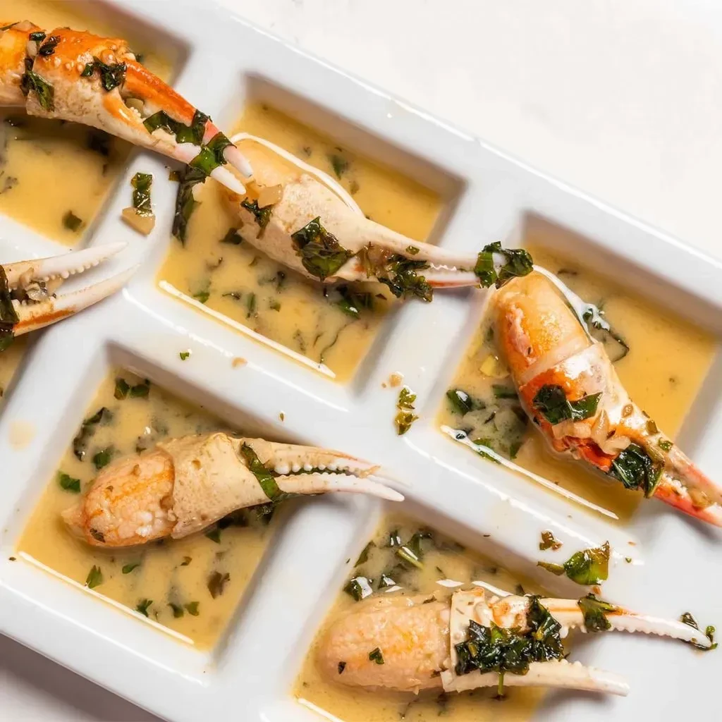 Six cooked crab claws with golden herb sauce in a partitioned dish.