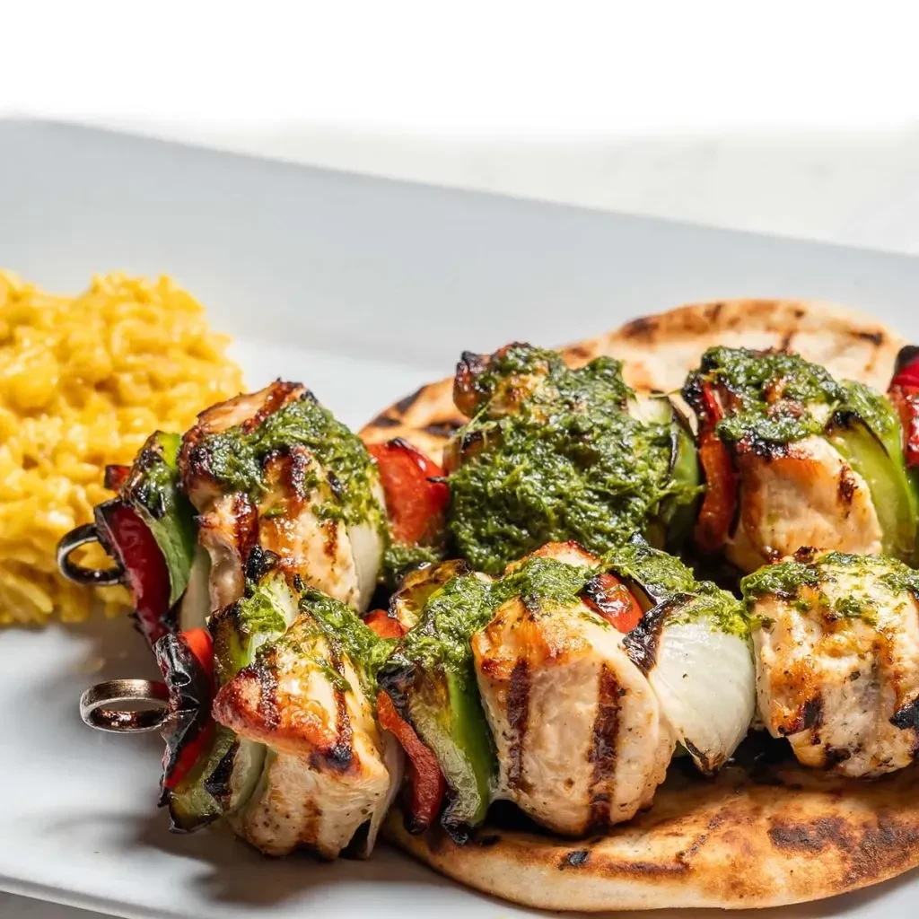 Grilled chicken and vegetable skewers with green sauce, yellow rice, and flatbread.