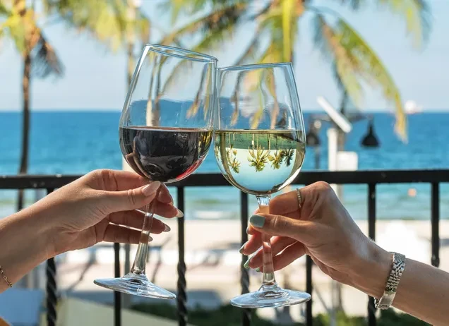 Two hands clinking red and white wine glasses, coastal background.