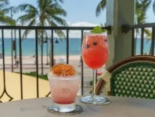 Two garnished cocktails on a table with a tropical beach and ocean view.