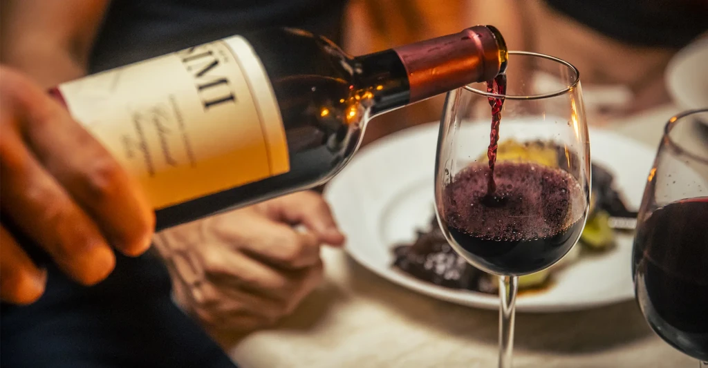 Red wine pours from a SIMI bottle into a glass, with food nearby.