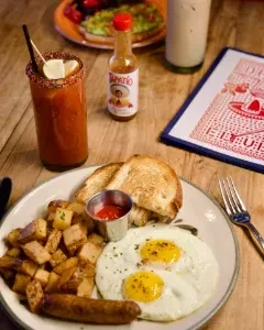 Breakfast plate of eggs, sausage, potatoes, hot sauce, and a spicy drink.