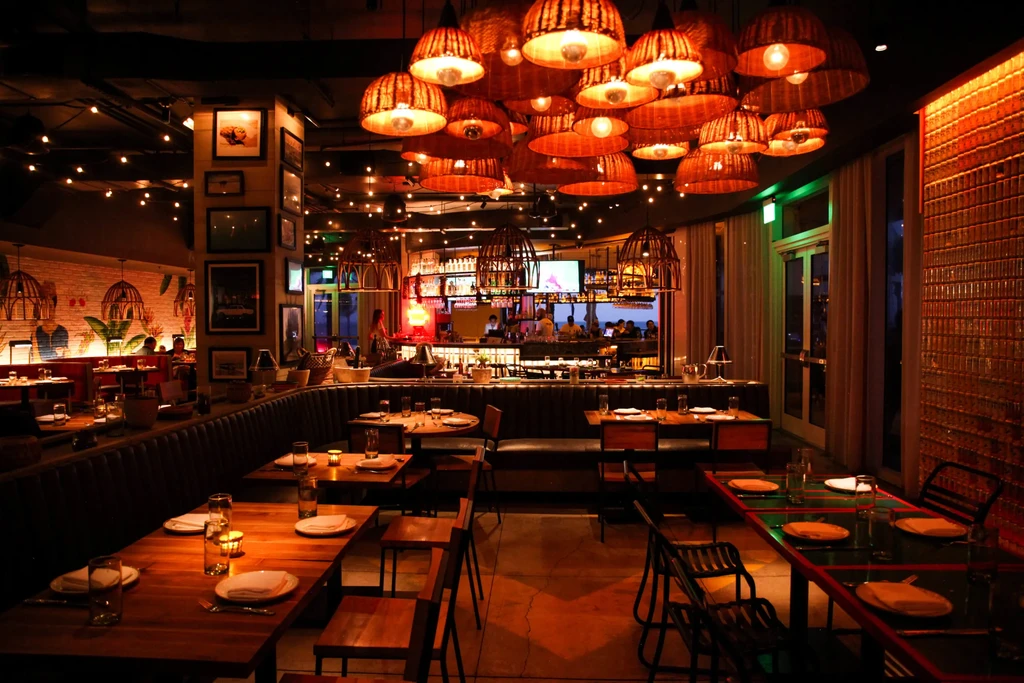 Dimly lit restaurant interior: wicker lamps, plush booths, set tables, lively bar.