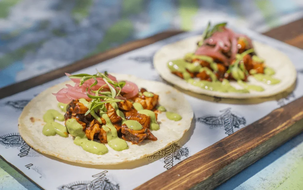 Two meat tacos topped with green sauce, pickled onions, and herbs.