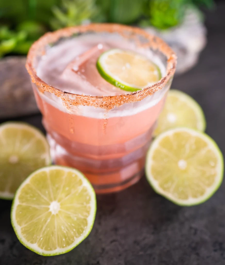 Pink cocktail in textured glass, spiced rim, with fresh lime slices.