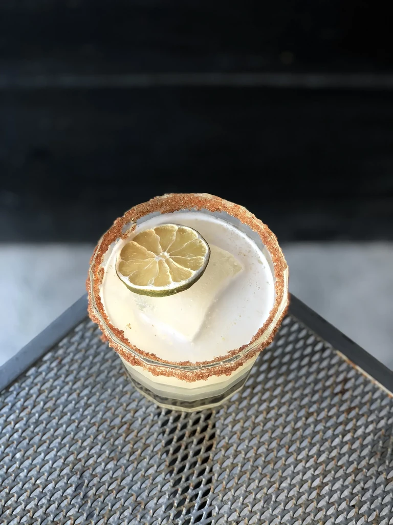 Faceted cocktail with spiced rim, dried lime, frothy liquid.