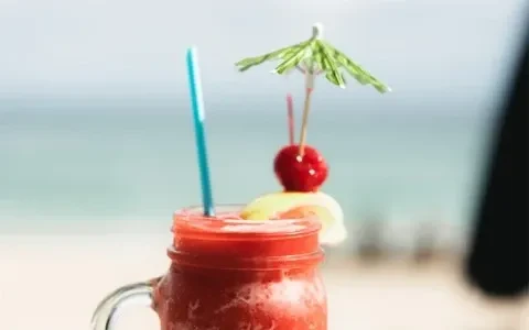 Red frozen drink in mason jar with garnishes, blurred beach background.