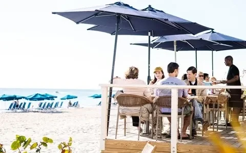 Restaurant deck with diners and server on a sunny white sand beach.