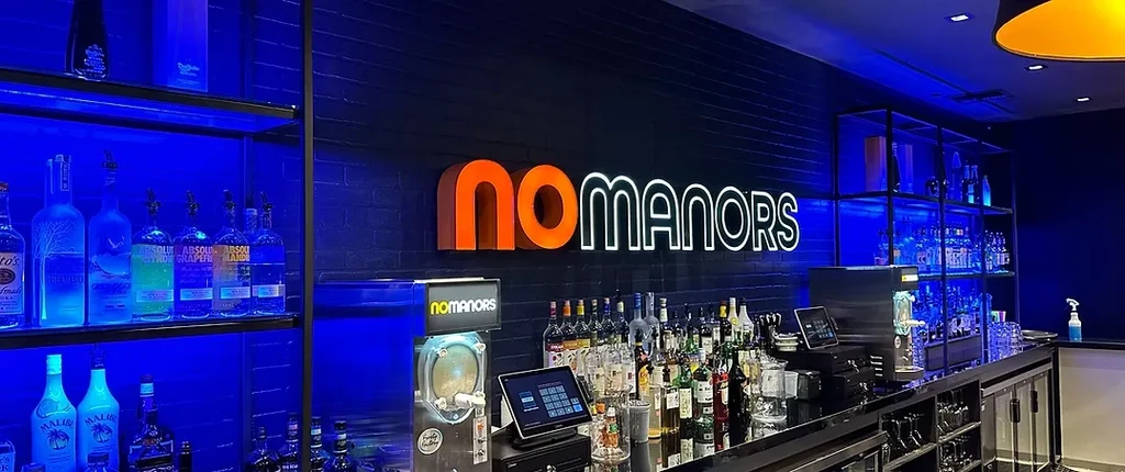 Blue-lit bar counter, glowing bottles, and 'no MANORS' sign.
