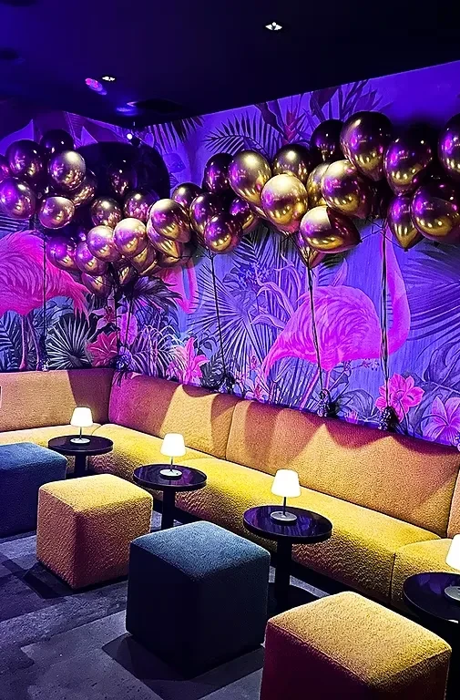 Purple-lit lounge with yellow sofa, flamingo wallpaper, and gold balloons.