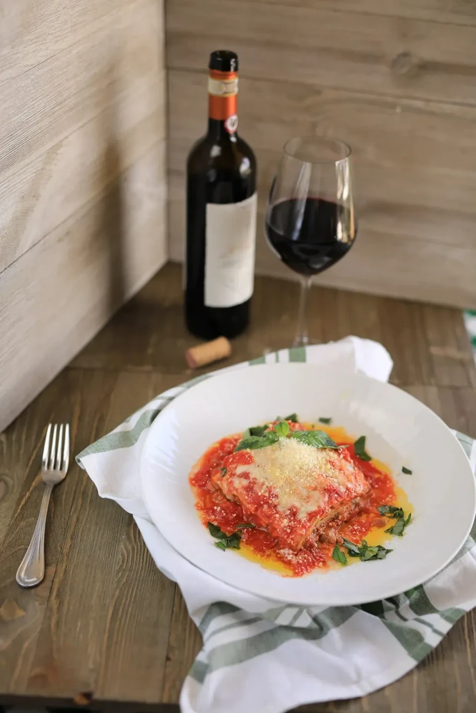 Plated lasagna or eggplant parmesan with sauce, cheese, basil, and wine.