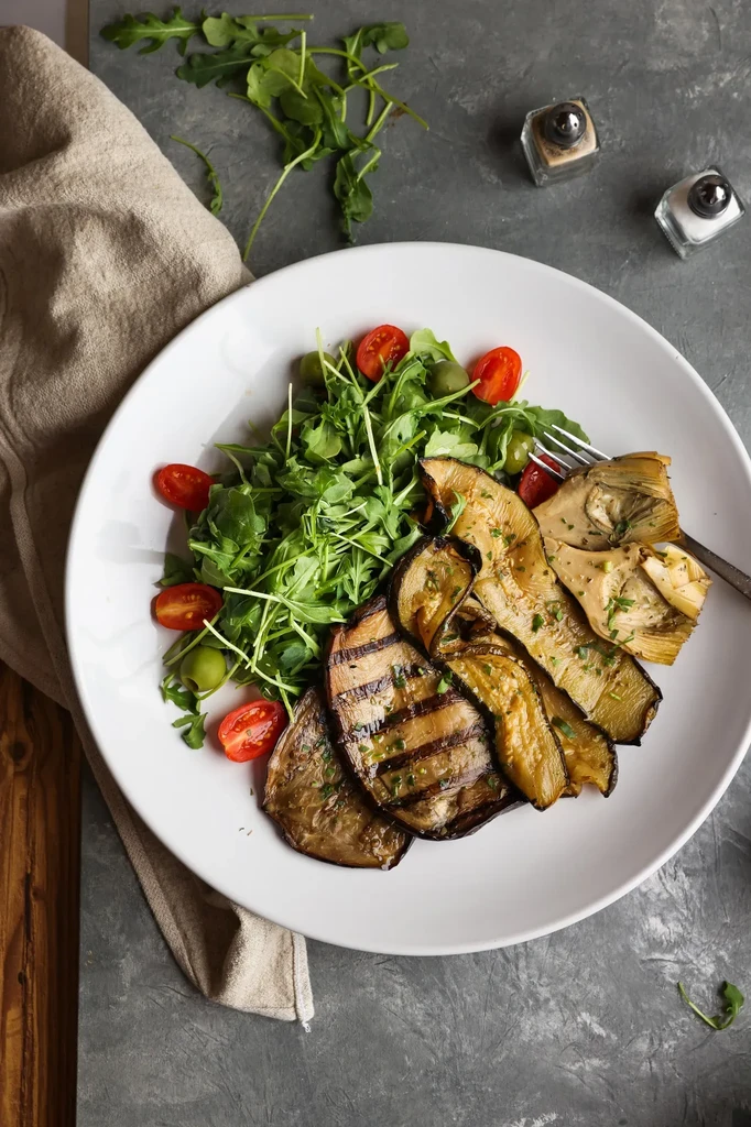 Grilled eggplant, artichoke hearts, and arugula salad on a white plate.