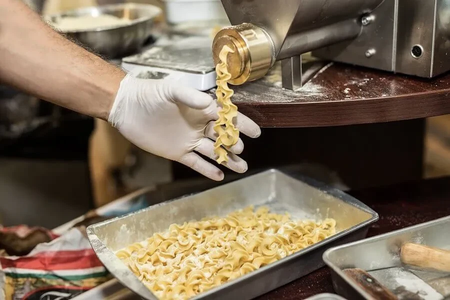 A gloved hand holds freshly extruded wavy pasta from a machine.