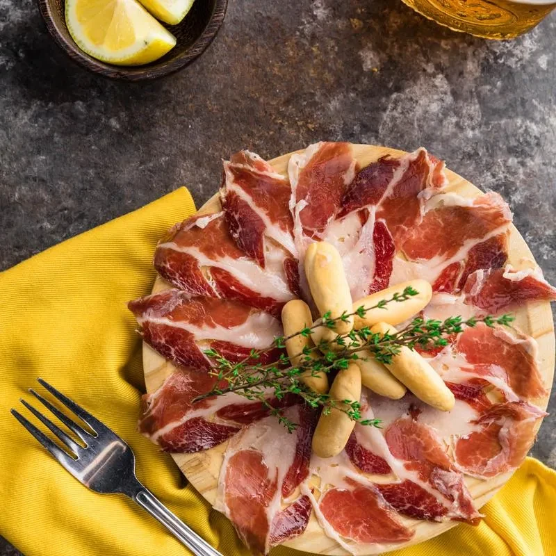 Overhead charcuterie board with cured ham, breadsticks, lemon, and beer.