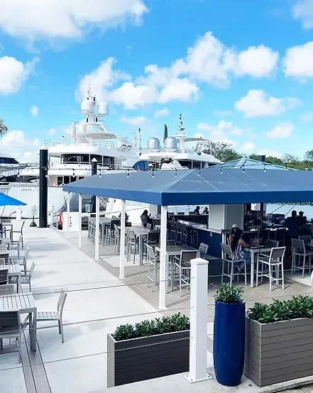 Waterfront dining with a blue awning, outdoor tables, and yachts.
