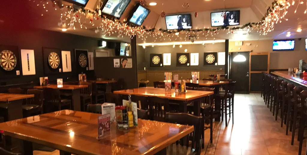 Dimly lit bar interior with high-top tables, a long bar, dartboards, and TVs.